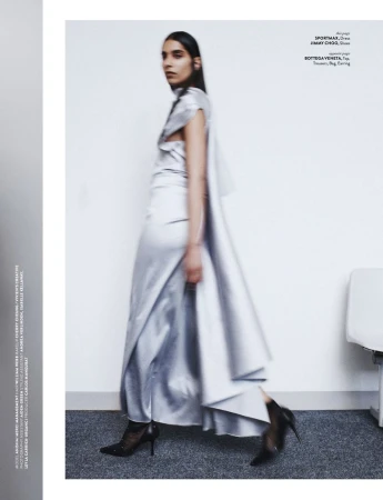 Editorial fashion shot: a woman in a silver dress and high heels walks against a white wall in a photo studio. The photo is blurred.