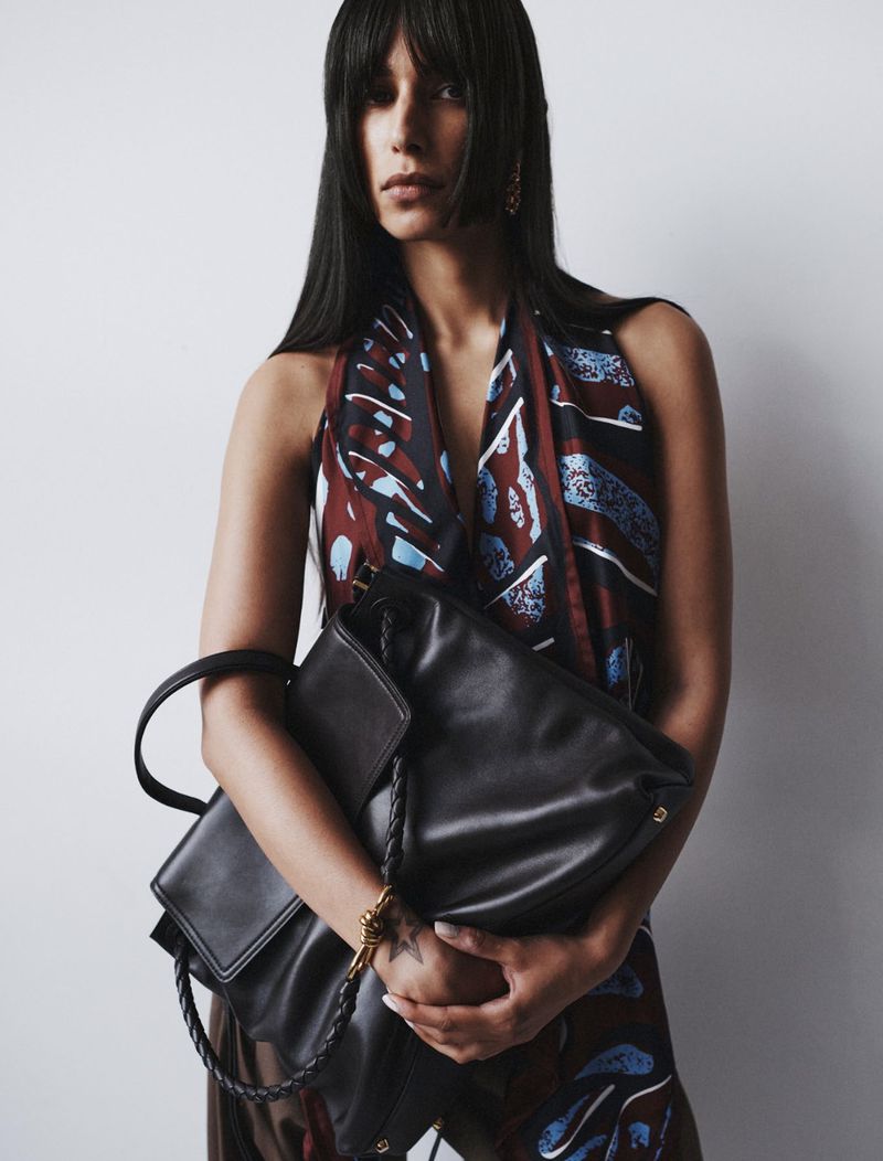 Portrait of a woman with dark hair and bangs holding a black leather handbag in a studio setting against a white background.