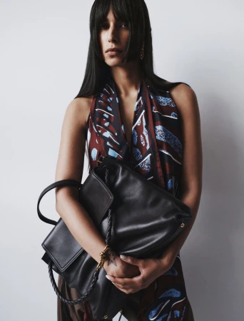 Portrait of a woman with dark hair and bangs holding a black leather handbag in a studio setting against a white background.