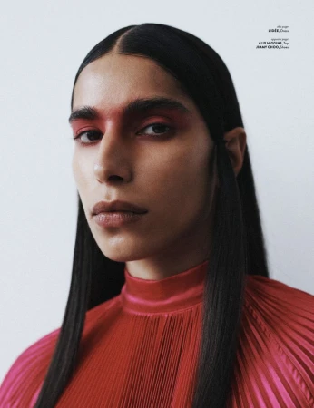 Close-up portrait of a slender adult with slick black hair and a striking red eyeshadow effect, wearing a red pleated top against a stark white background.