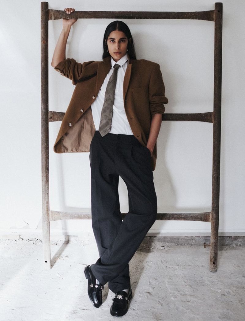 Adult posing in a photo studio in a brown blazer, white shirt, dark tie, dark trousers, and black loafers near a ladder.