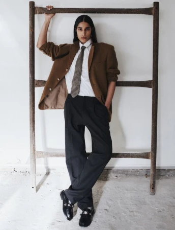 Model Anisha Sandhu poses in an oversized brown blazer, necktie, and black trousers leaning on a metal frame against a white wall.
