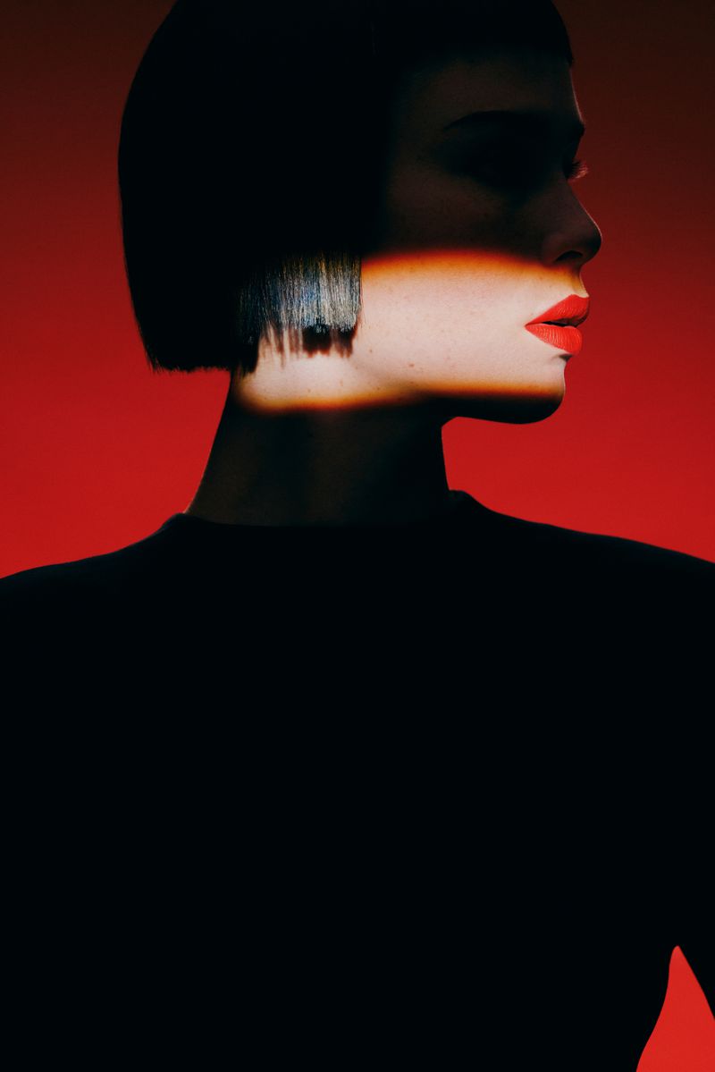 Portrait of a young woman with a bob haircut and red lipstick against a red background. A beam of light crosses her face.