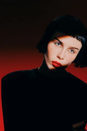 Portrait of a stylish adult female with short dark hair and bold red lipstick, illuminated by a beam of light against a red background.