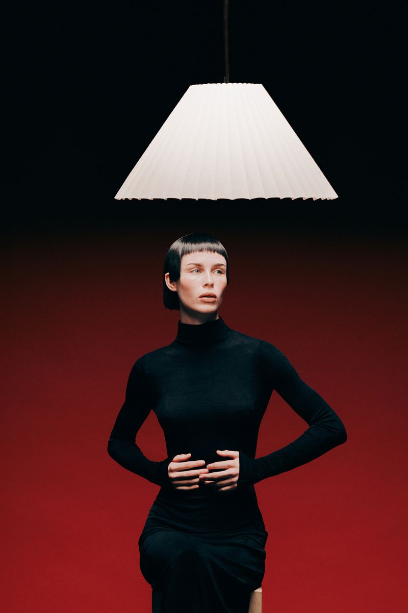 Woman in black turtleneck dress seated under a pleated white lamp on a red and black gradient backdrop in a studio setting.