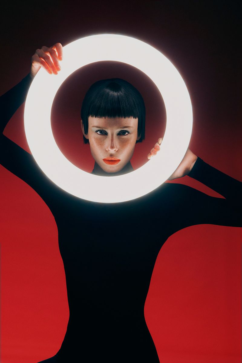 Woman in black bodysuit holding glowing ring in front of her face, looking at camera, against red background in studio setting.