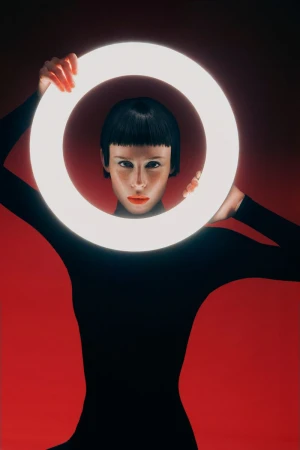 Woman in black bodysuit holding glowing ring in front of her face, looking at camera, against red background in studio setting.