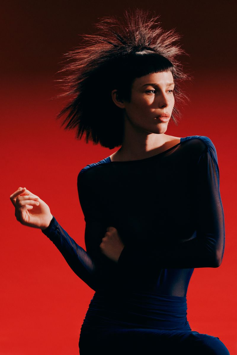 Model Jesi Evans with spiky black hair in a sheer navy long-sleeve garment posing against a vivid red backdrop.