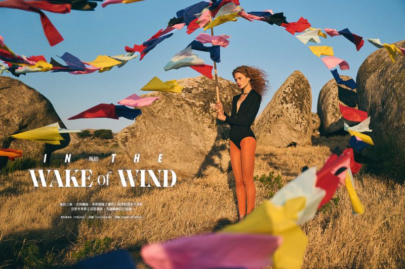 A fashion shot in a dry, rural outdoor setting. A model in a black bodysuit poses amidst rocks and colorful flags under a clear blue sky.