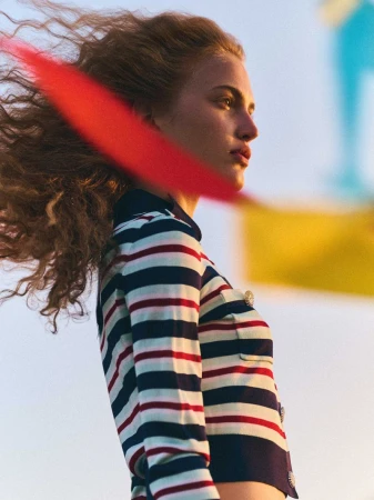 Woman with windblown curly hair, wearing a striped collared shirt, with blurred flag shapes in the background, low angle, outdoor.
