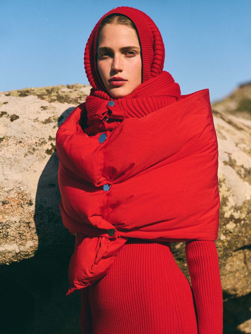 Outdoor fashion portrait of a young adult female wearing a red knit dress and a red puffer scarf with blue buttons against a blue sky.