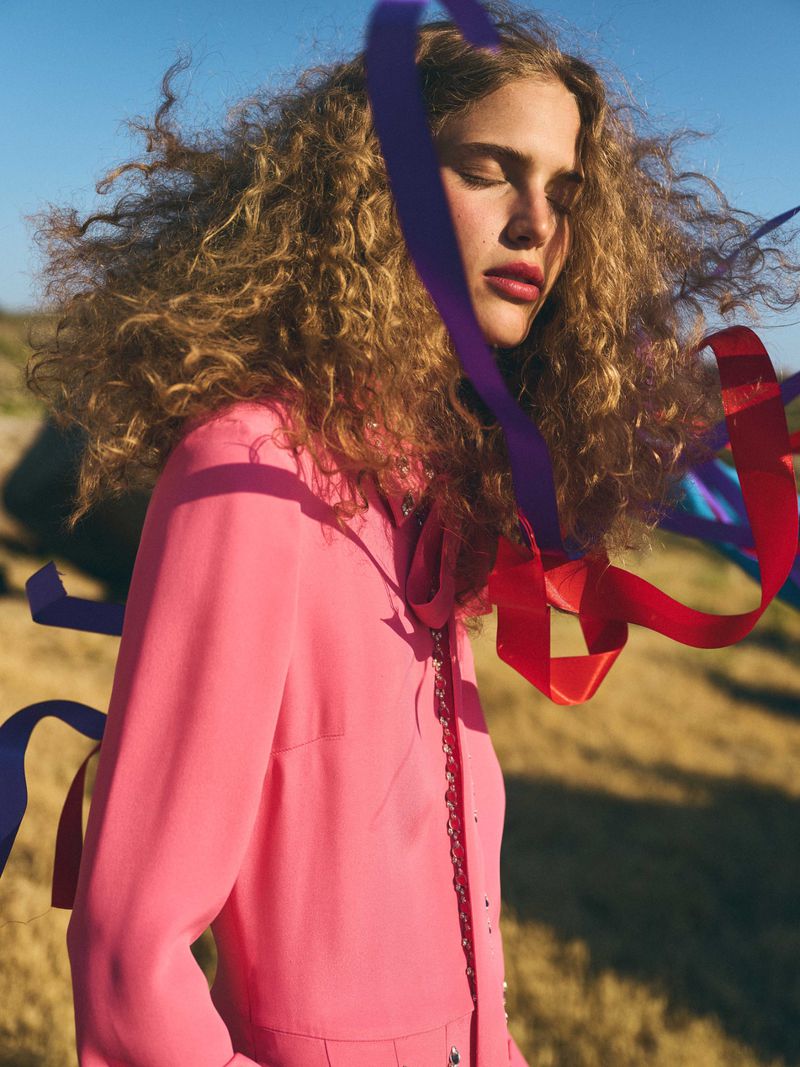A young adult woman with curly hair poses outdoors in a pink top, with ribbons and a blue sky backdrop, showcasing a fashion editorial.