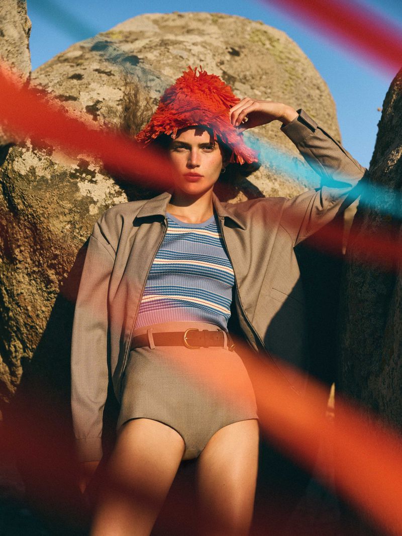 Woman wearing a striped shirt, jacket, high-waisted shorts, and fringed hat posing outdoors amongst rocks in a fashion style portrait.