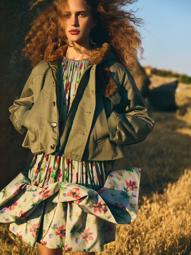 Fashion shot of a woman with curly hair wearing a floral dress and an olive green jacket posing in a field during the daytime.