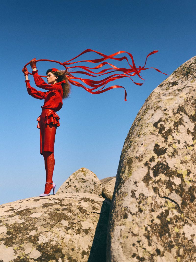 Fashion editorial shot of a woman in a red outfit holding a red ribbon while posing on a rocky landscape against a blue sky background.