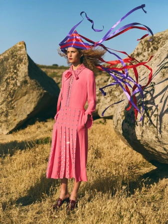 A young adult female fashion model stands outdoors in a pink dress and headpiece with flowing ribbons, posing in a field of dry grass.