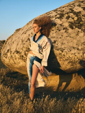Fashion shot of a young adult female model with curly red hair, wearing a kimono-style outfit, posing beside a massive boulder in a field.
