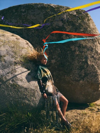 A fashion shot outdoors shows a young adult woman with ribbons, wearing a unique dress, leaning against a textured rock formation on a sunny day.