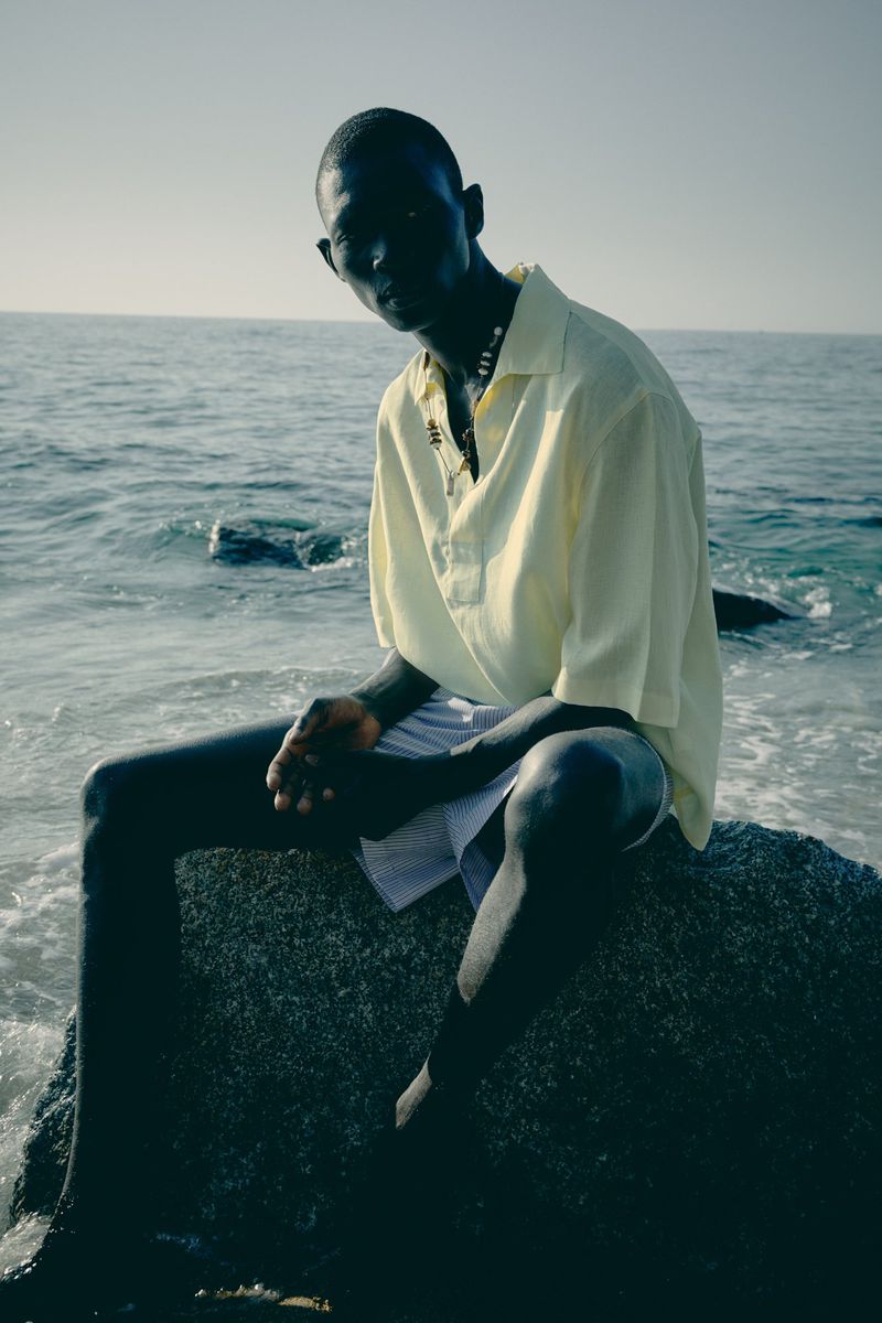 Model Fernando Cabral sitting on a dark ocean rock wearing a light yellow polo shirt and shorts by the sea.