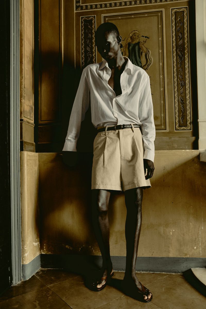 Adult male fashion model in casual wear poses in an ornate corner with architectural trim, neutral colors, and soft, diffused lighting.