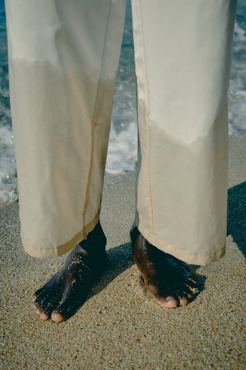 Close-up of a person's bare feet standing on the beach sand, with ocean waves gently lapping at the shoreline and beige pants.