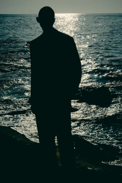 Silhouette of an adult male in a suit standing at the ocean's edge, with the sun reflecting on the water in the background.