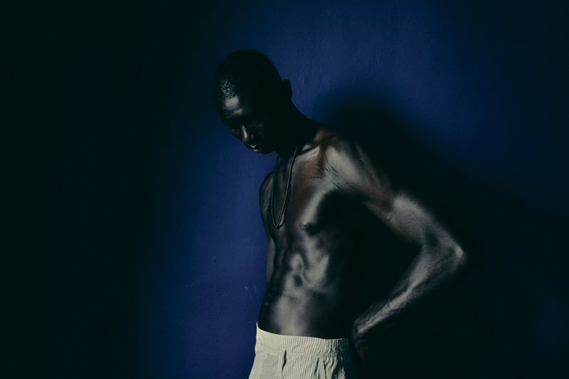 A shirtless man with dark skin stands against a dark blue wall. His head is tilted down, and he wears light-colored pants and a necklace.