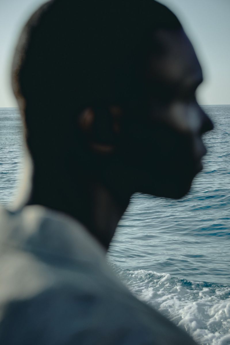 Profile of a blurred Black person by the ocean, featuring sea waves, blue water, and clear sky in a summertime outdoor setting.