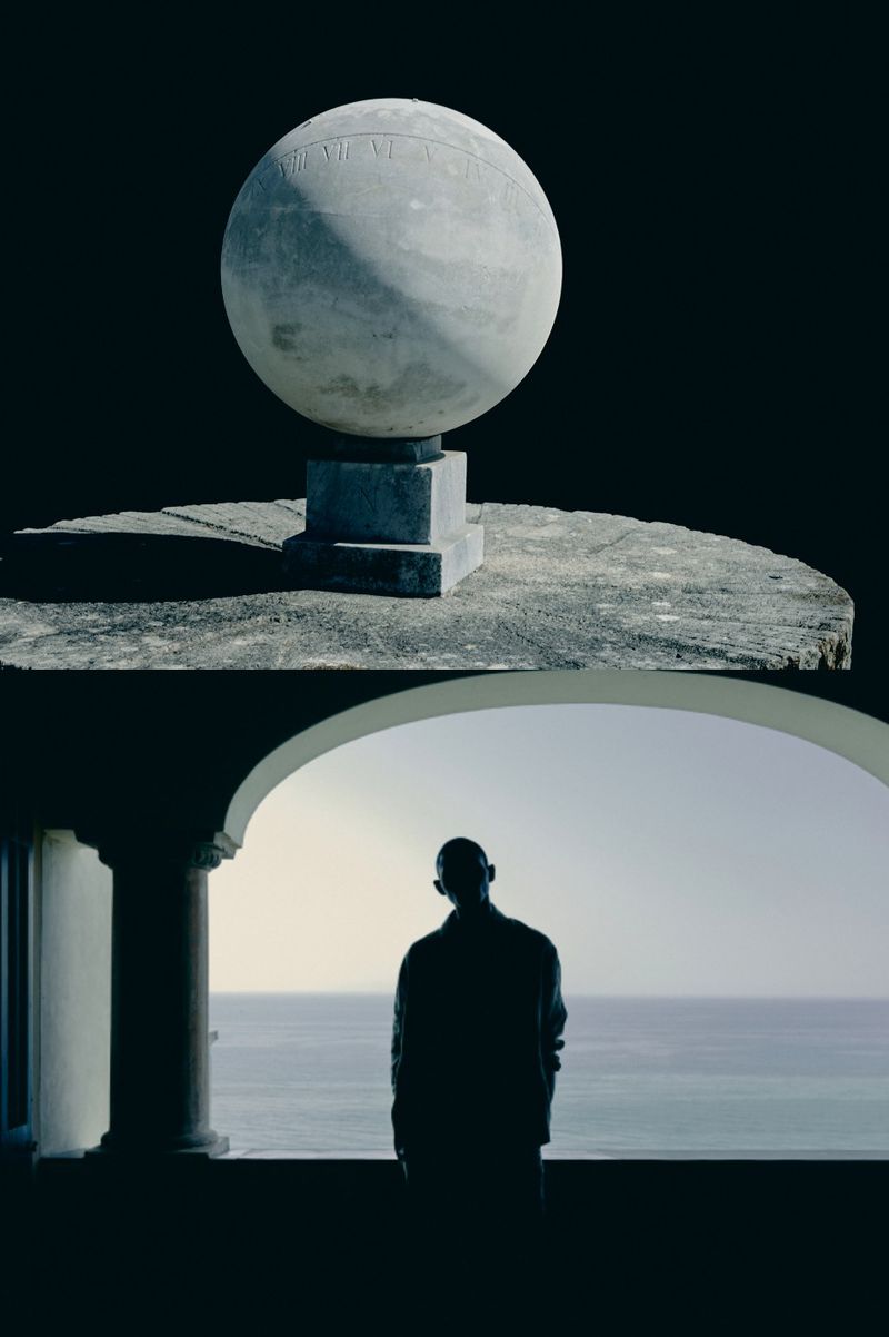 Architectural image of a dark silhouette of a person under an arch with a sea view and a spherical marble statue above.