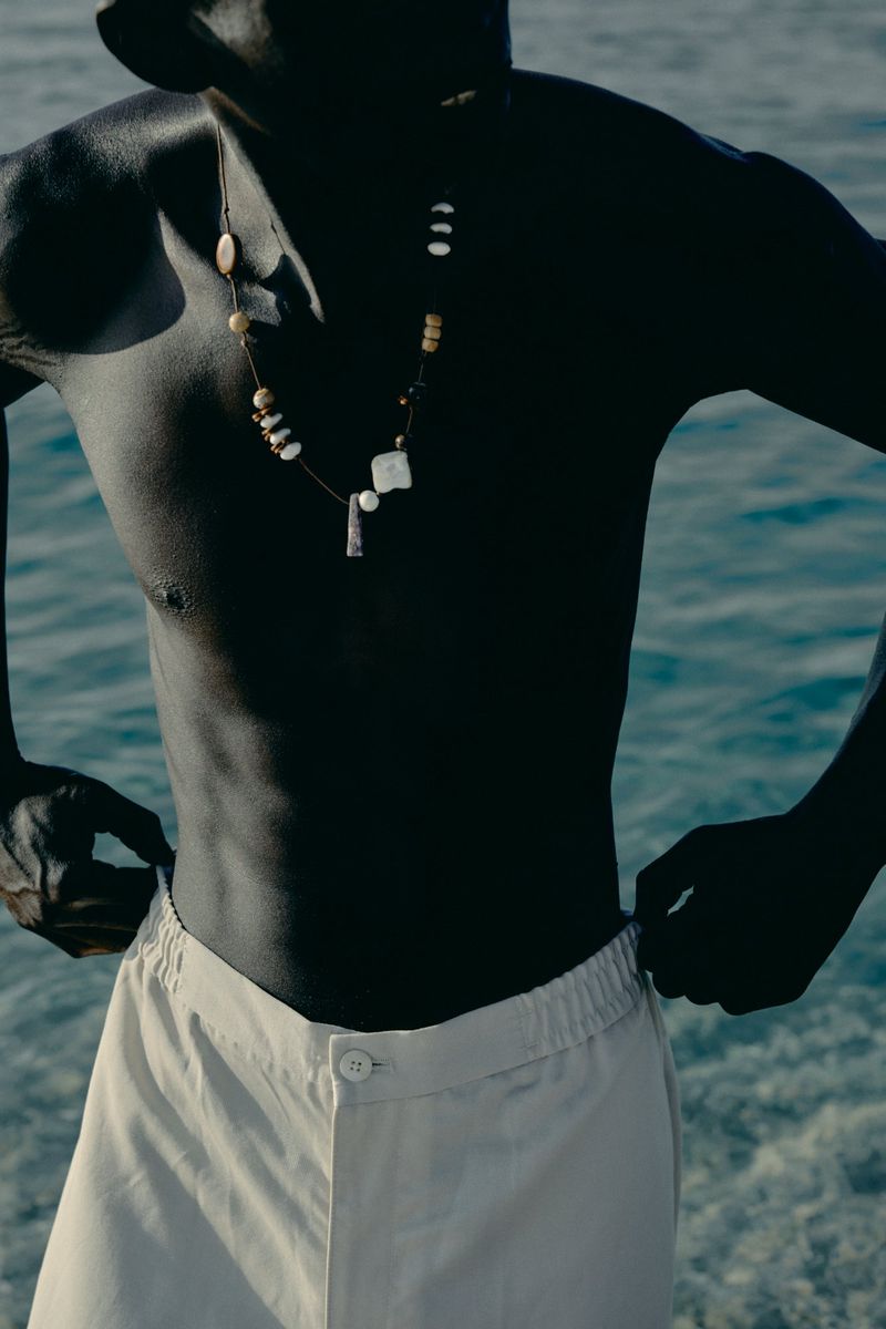 Portrait of a dark-skinned adult man wearing light-colored pants and a stone beaded necklace against a rippled water background.