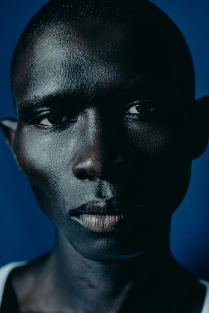 Portrait of a Black male, close-up shot with dark skin and intense gaze, set against a dark blue background, showcasing details and textures.