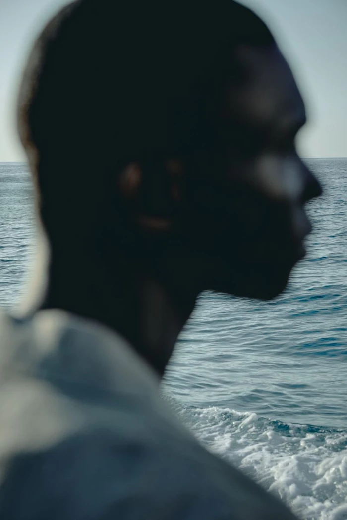 Close-up profile of a black man in silhouette against an ocean background with choppy blue water.