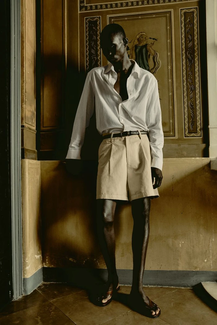 Full-length portrait of a male model wearing a white shirt and beige shorts against an ornate classical interior wall.