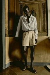 Full-length portrait of a male model wearing a white shirt and beige shorts against an ornate classical interior wall.