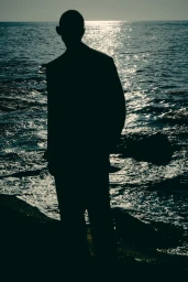 Silhouette of model Fernando Cabral in a tailored suit standing at the water's edge against glittering ocean waves.