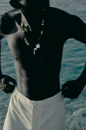 Model Fernando Cabral standing shirtless wearing a beaded necklace and off-white trousers against a blue water background.