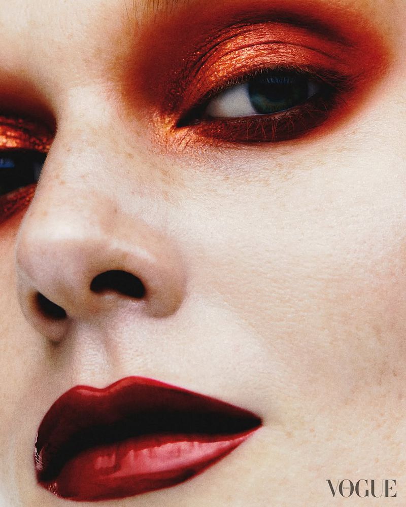 Close-up portrait of a woman's face with bold makeup, including shimmering red eyeshadow and glossy red lipstick, and 'Vogue' logo.