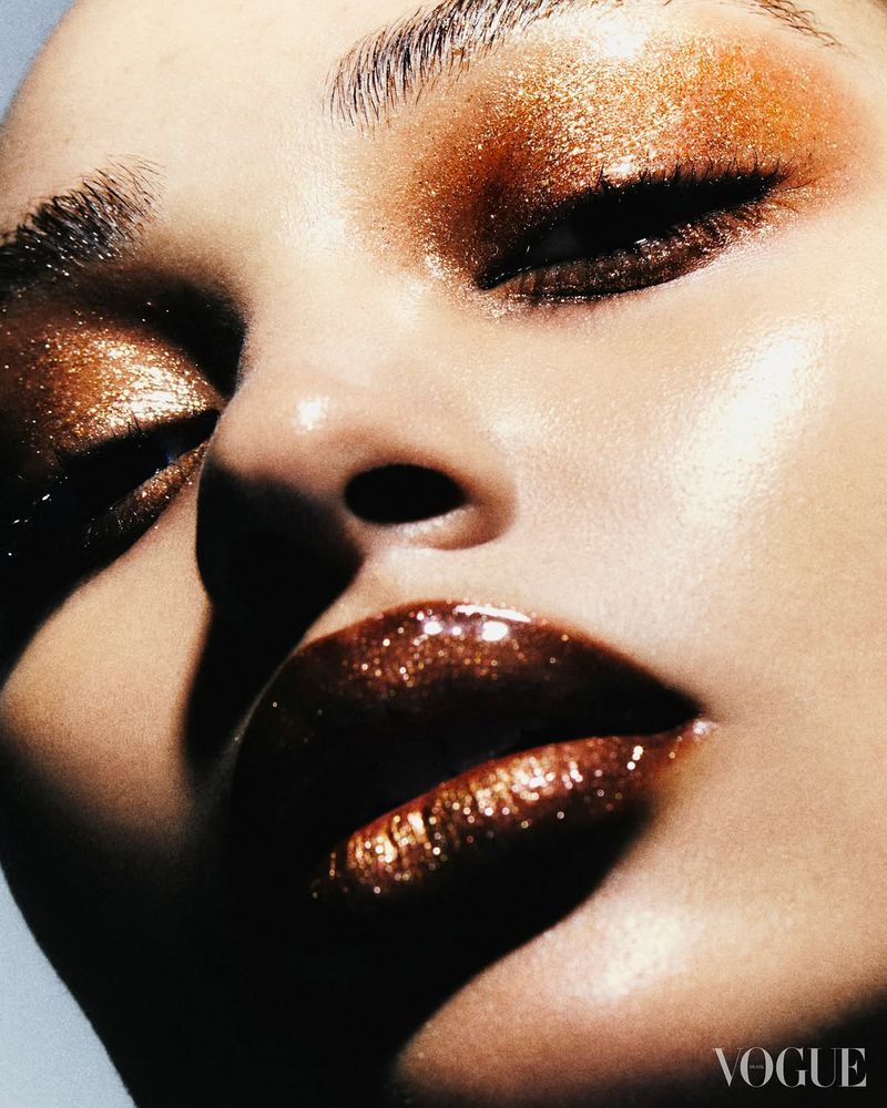 Close-up of a woman's face with bronze glitter eyeshadow and glossy lipstick with studio lighting in a beauty editorial.