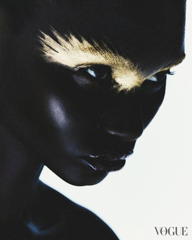Striking close-up portrait of a dark-skinned woman with golden makeup on her brow, showcasing dramatic beauty against a white backdrop.