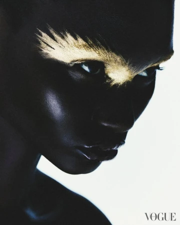 Striking close-up portrait of a dark-skinned woman with golden makeup on her brow, showcasing dramatic beauty against a white backdrop.