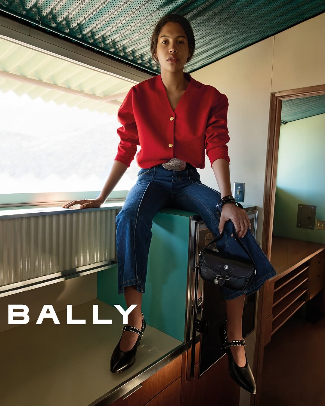 Model wearing a red cardigan and denim jeans seated indoors holding a black handbag for Bally SS25 campaign.