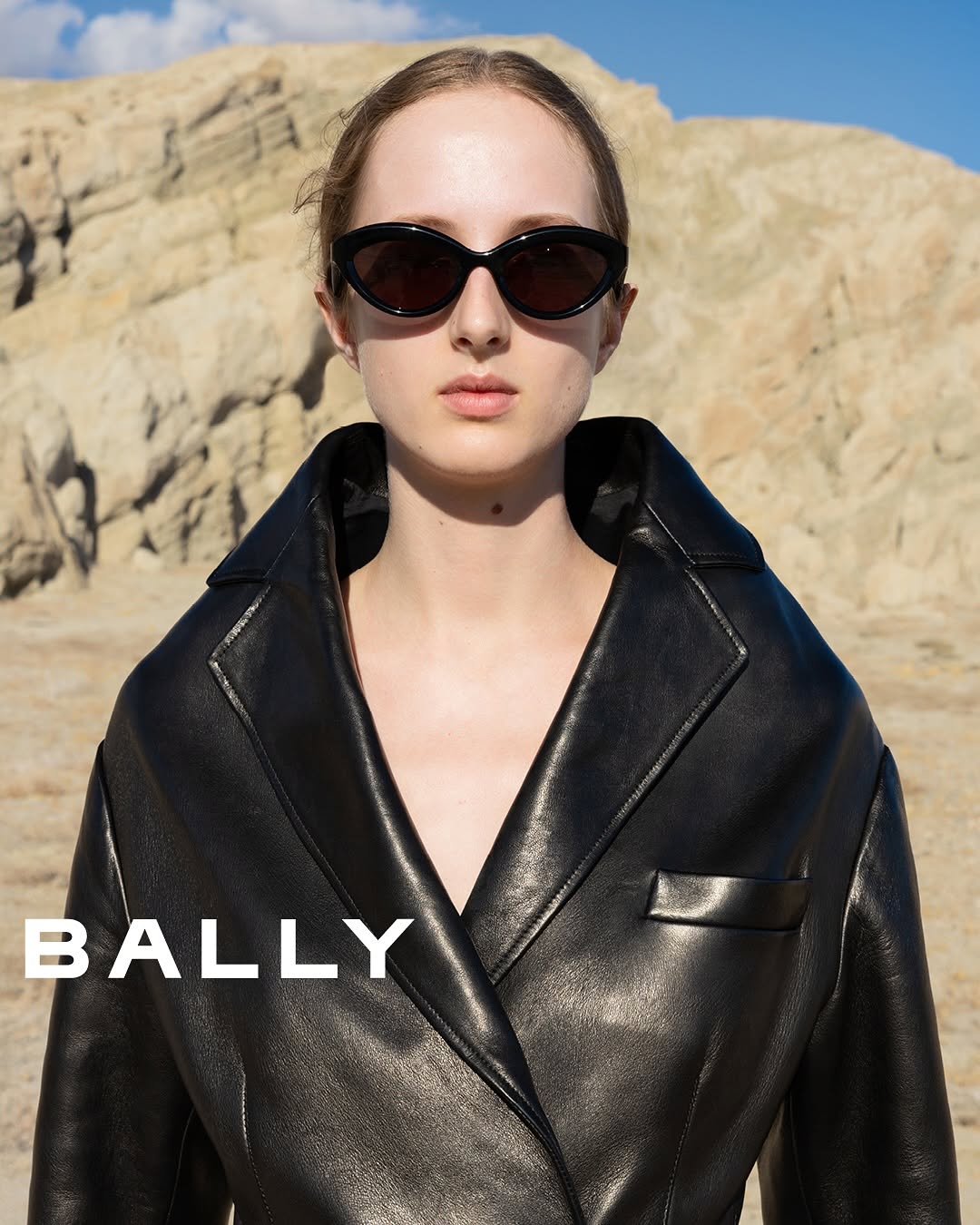 Fashion shot of a young woman in sunglasses and a black leather jacket, posing outdoors against a desert-like landscape under blue sky.