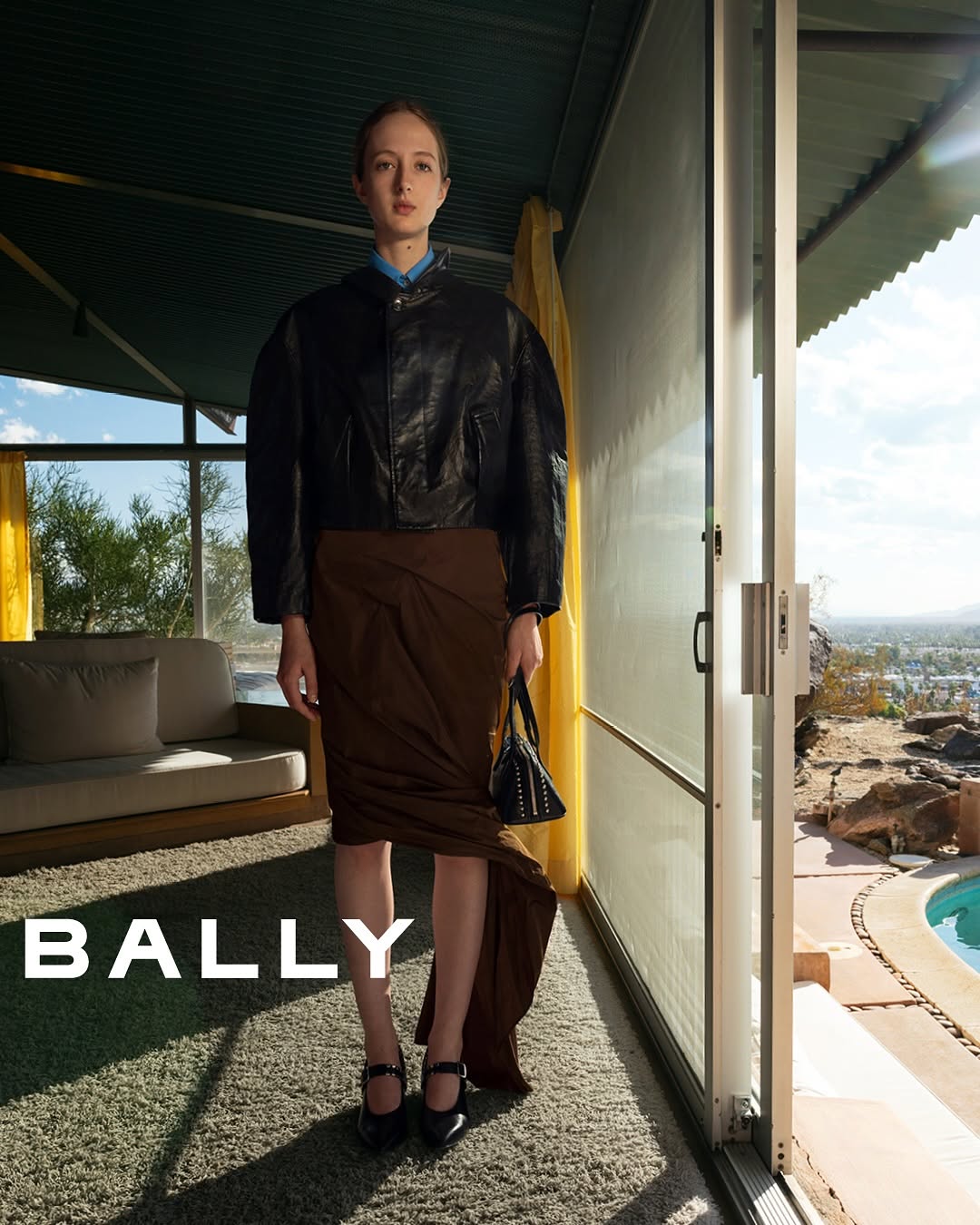 A slender woman models a black leather jacket and draped brown skirt in front of a modern home interior and pool. Fashion shot.
