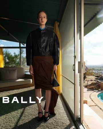 A slender woman models a black leather jacket and draped brown skirt in front of a modern home interior and pool. Fashion shot.