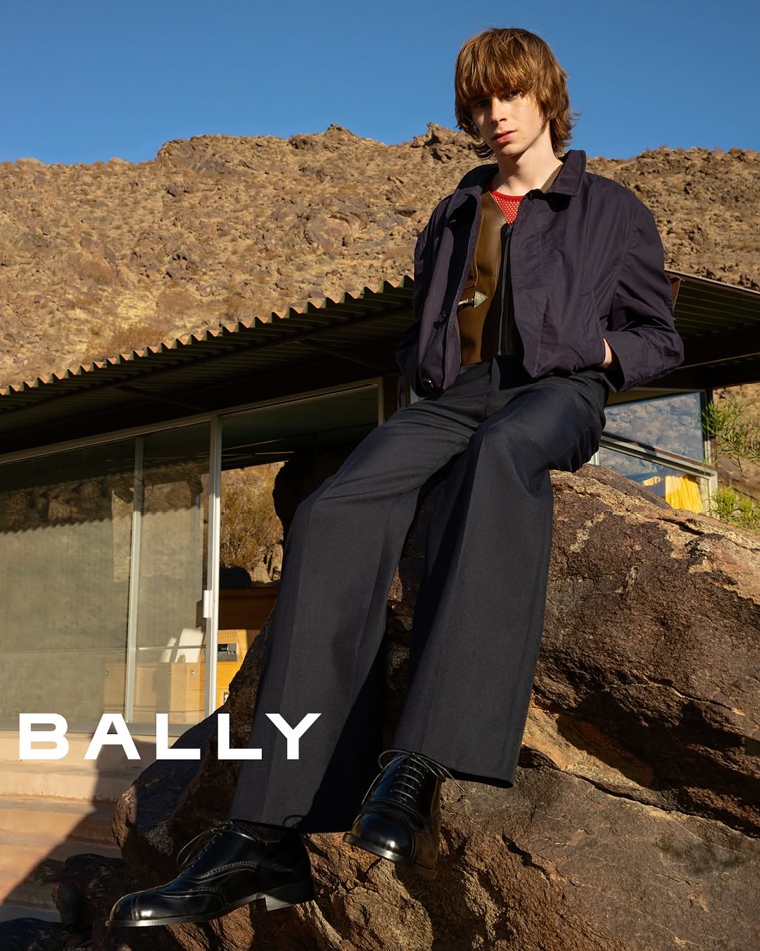 An adult male model sits on a rock wearing a dark jacket and trousers, showcasing Bally shoes, against a desert mountain backdrop.