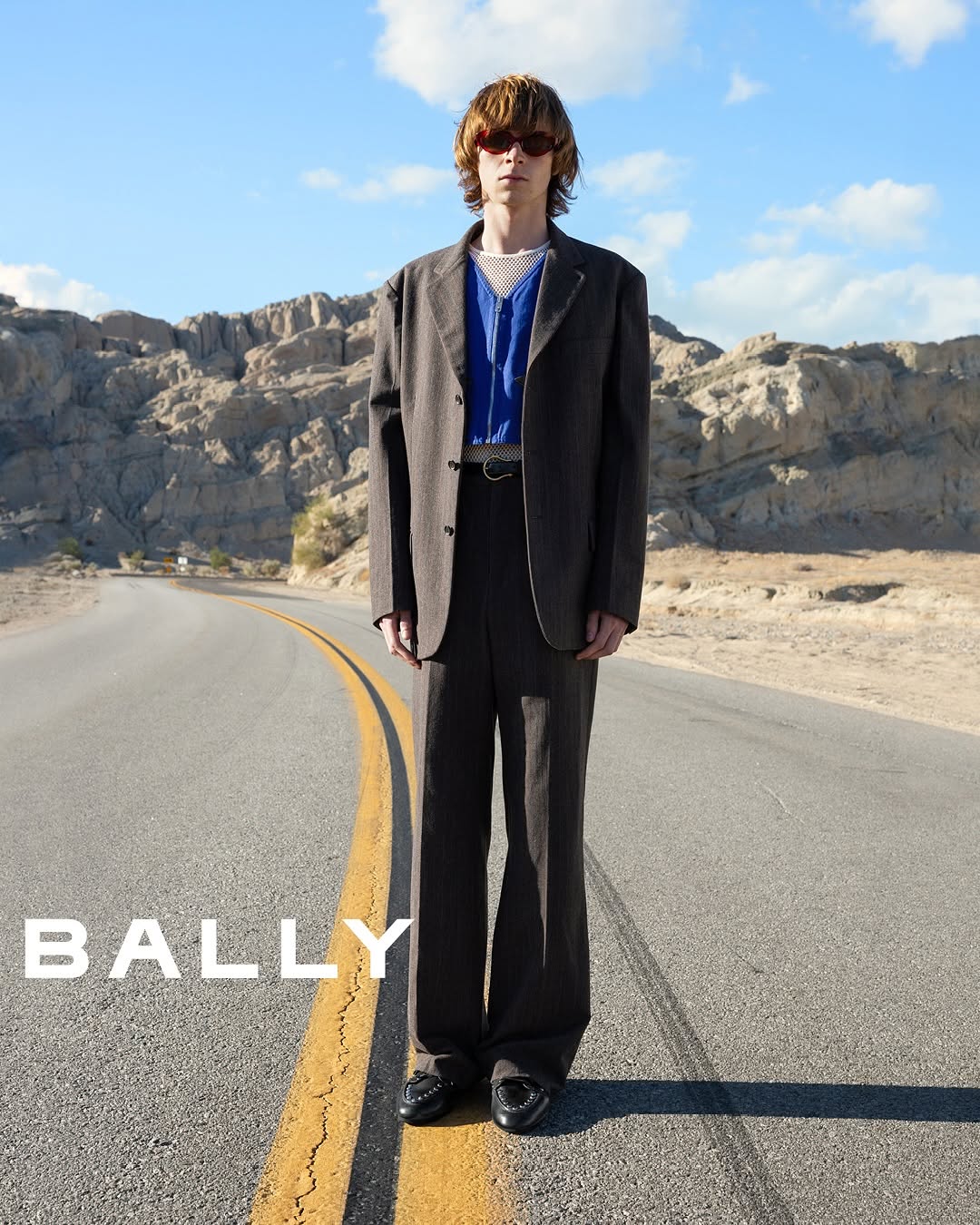 Fashion model in a suit posing on a desert road with distant mountains under a blue sky for Bally, showcasing style and landscape.