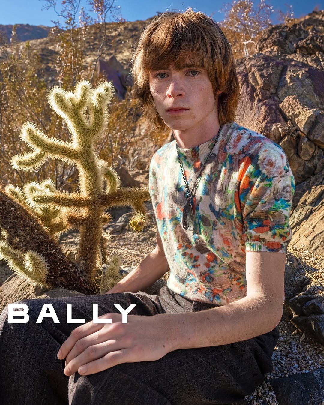 Adult male poses in a desert environment next to a cactus while wearing a floral short-sleeve shirt and pants. Bally logo is visible.