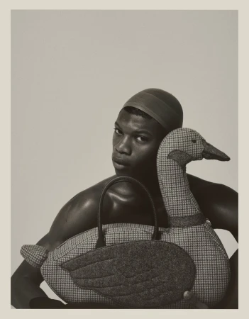 Model Razak Salifou poses shirtless holding a large houndstooth fabric duck-shaped handbag against a neutral studio backdrop.