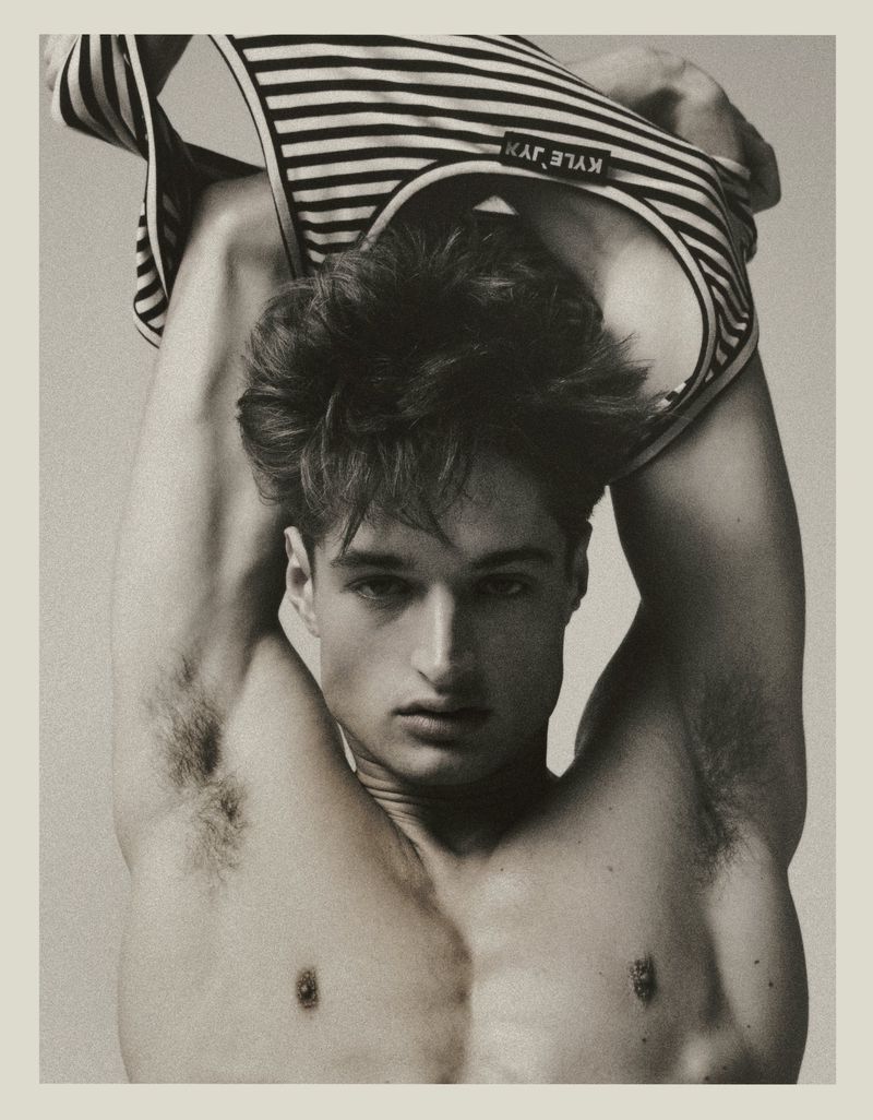 Monochrome portrait of a shirtless adult male model pulling a horizontally striped tank top over his head in a photo studio.