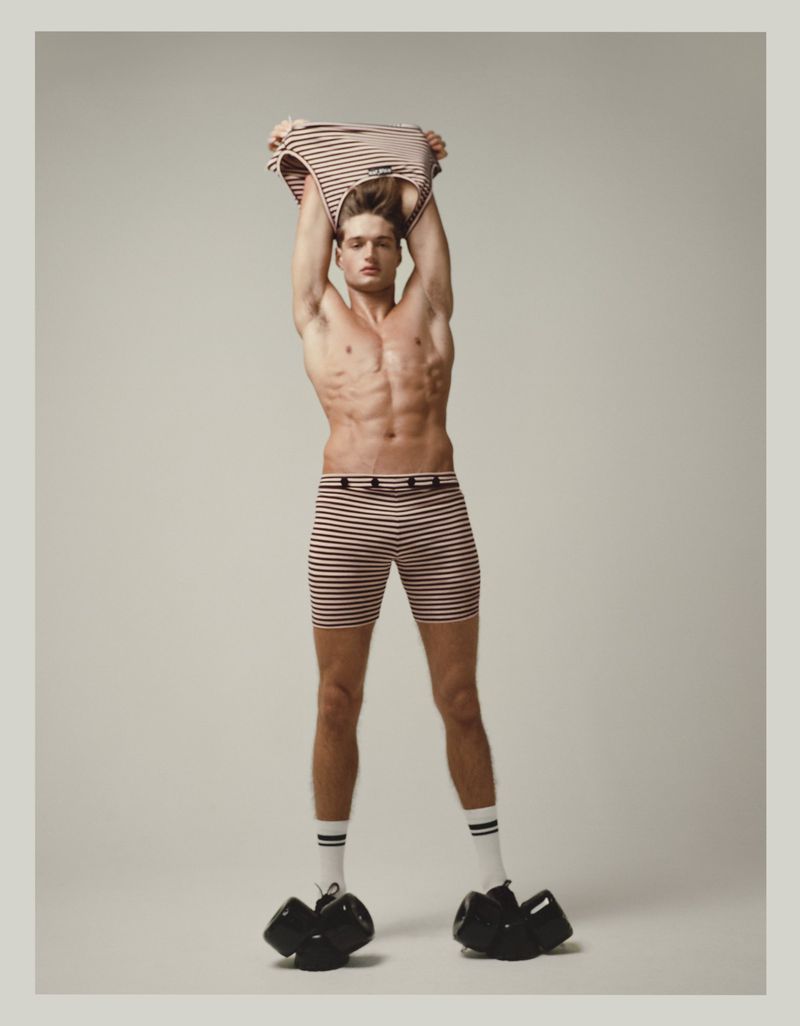 Athletic man in striped shorts and socks lifts matching shirt overhead against a neutral backdrop in a fitness-themed studio shot.
